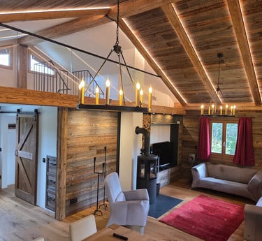 Großzügiges Wohnzimmer mit offenem Altholz-Dachstuhl, Galerie mit Glasgeländer, zwei Kronleuchtern und Kaminofen.