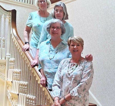 Four smiling senior women posing together on a historic wooden staircase in a bright home.