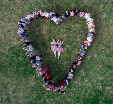 Hochzeitsgesellschaft formt Herz auf Wiese um liegendes Brautpaar. Drohnenaufnahme von Joel Pingel.