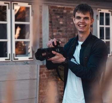 Lächelnder Hochzeitsfotograf Joel Pingel mit Kamera vor Backsteingebäude. Portrait: Just Perspectives.