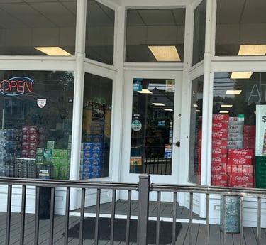 Entrance of Round Hill Market with an "OPEN" sign, lottery ad, and soda boxes visible through the glass doors.
