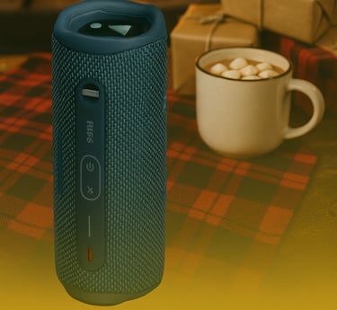 JBL Flip 6 Bluetooth speaker on plaid blanket with hot cocoa and wrapped gifts by a fireplace.