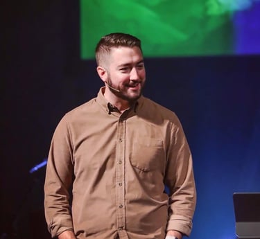 Cole speaking on stage at a ministry event, wearing a tan shirt with a lapel microphone.
