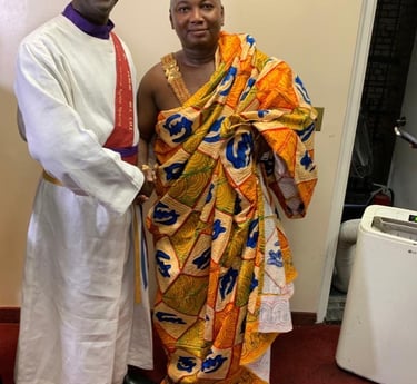 A smiling priest in a white alb standing next to a man wearing a traditional colorful Ghanaian Kente cloth.