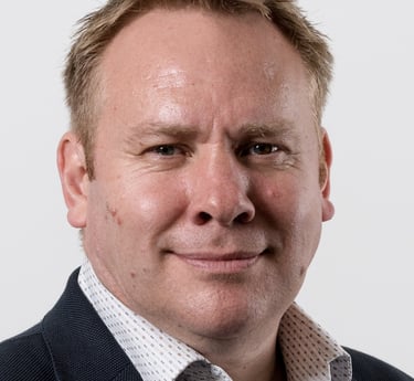 Professional headshot of a middle-aged man with short blonde hair wearing a suit jacket and collared shirt.