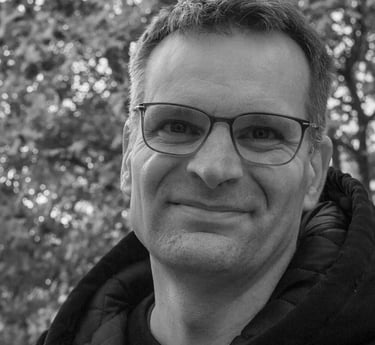 Black and white portrait of a smiling middle-aged man with glasses outdoors against bokeh trees.