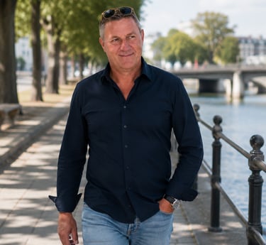A middle-aged man in a navy shirt and jeans walking along a riverside path with sunglasses.