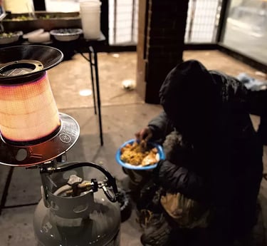 Man sitting on the ground eating his plate of food in front of a propane heater.