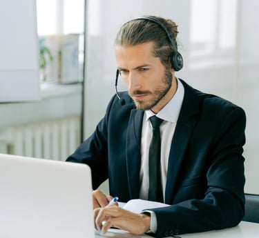 A man with a beard and long hair tied back is wearing a suit and headset while working on a laptop. He holds a pen and appear