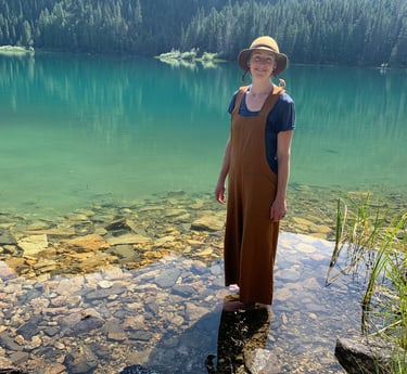 woman with hat standing in green water. rocks visible in water and mountain in background