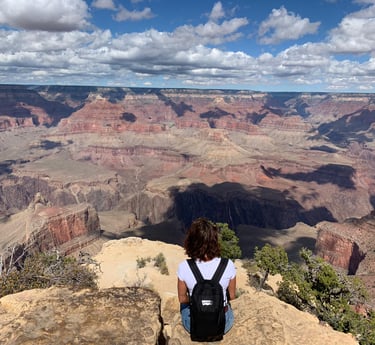 A hiker with a backpack sits on a rocky cliff overlooking the vast Grand Canyon landscape under a cloudy blue sky.