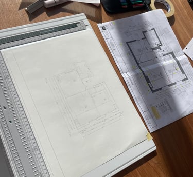 Architectural floor plan sketches and blueprints on a wooden desk with a drafting board and tools.
