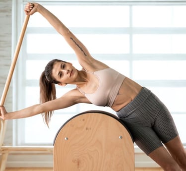 A woman performs a side stretch on a wooden Pilates small barrel with a pole for core stability.