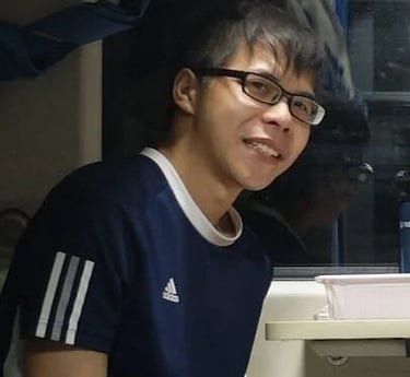 Smiling young Asian man wearing glasses and a blue Adidas t-shirt sitting indoors.