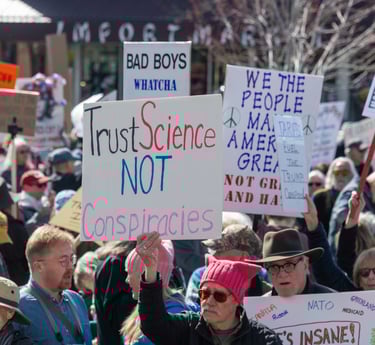 Protesters at a political rally holding a Trust Science Not Conspiracies sign among a diverse crowd.