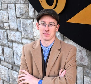Professional man wearing a tan overcoat, flat cap, and glasses posing against a stone wall.