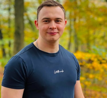 A smiling man in a navy blue t-shirt stands outdoors against a background of yellow autumn foliage.