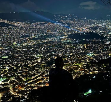 Silhouette of a person overlooking the illuminated city skyline of Medellin at night from a mountain viewpoint.