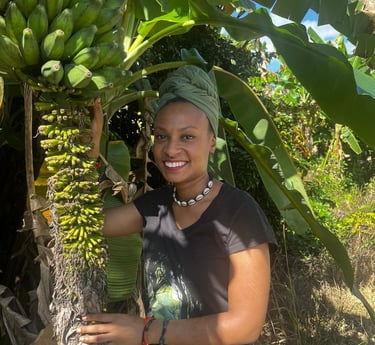 Evvie, a brown-skinned black femme person in a green head-wrap stands underneath a banana tree