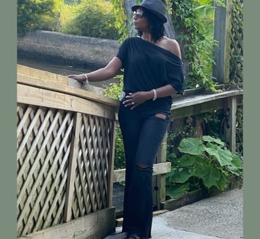 Woman in stylish black off-the-shoulder top and fedora posing on a wooden bridge outdoors.