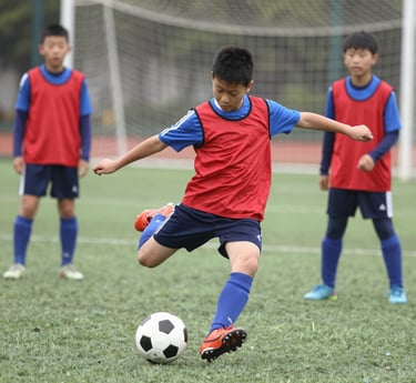 A group of young female athletes in sports attire are gathered together, some wearing red jackets or maroon jerseys with the logo of a soccer academy. They appear to be standing in front of a wire fence with spectators in the background, dressed in casual clothing.