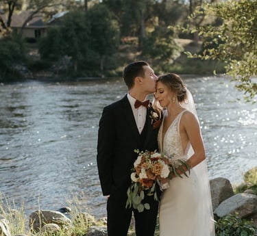 Groom kisses bride on the forehead during a scenic outdoor riverside wedding ceremony.