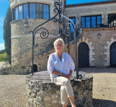 A woman sits on an old stone well in front of a historic European stone building with a round tower.
