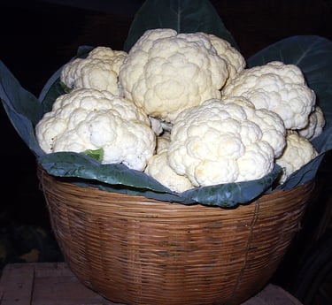 Fresh NXG DELIGHT white cauliflower heads with green leaves in a woven wicker basket at a market.