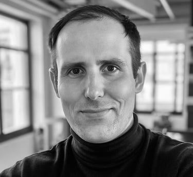 Black and white professional headshot of a smiling man wearing a black turtleneck in a bright studio office.