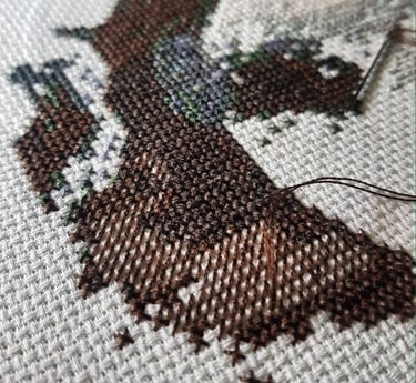 Primer plano de hilo de bordar marrón y verde sobre tela blanca para punto de cruz, con una aguja.