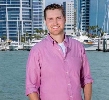 Josiah Ryan, business consultant wearing a pink shirt and jeans standing in front of a body of water