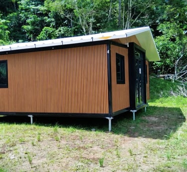 A modern prefabricated tiny house with wood-look siding and a white roof in a lush tropical garden.