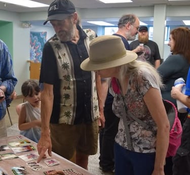 A group of people gathering around a table to look at a collection of small art cards at a community event.