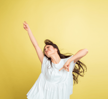 L'immagine mostra una donna che indossa una blusa bianca, posando con un'espressione energica e gioiosa su uno sfondo giallo.