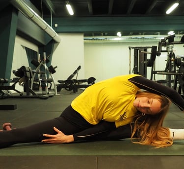 Una donna su un tappetino da yoga in palestra esegue una posa laterale di stretching, tenendo il piede con una mano mentre si