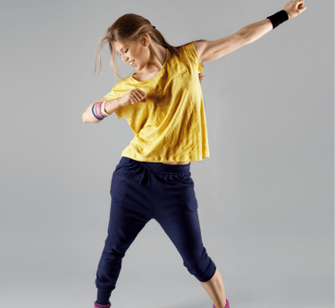 Una donna che danza con energia, indossando una maglietta gialla, pantaloni blu scuro e scarpe sportive nere con dettagli tur