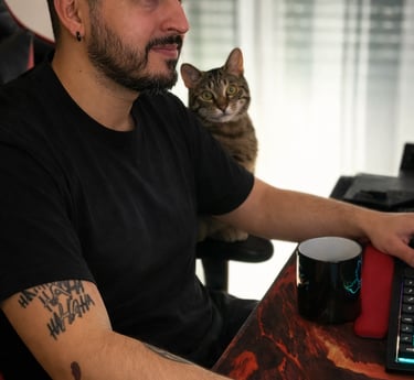 Bearded man with tattoos gaming at a desk while a tabby cat sits on his shoulder.