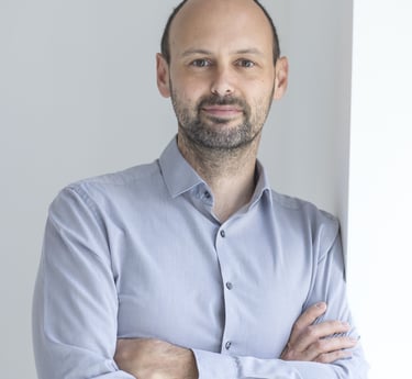 Professional headshot of a smiling man with a beard wearing a grey button-down shirt with arms crossed.