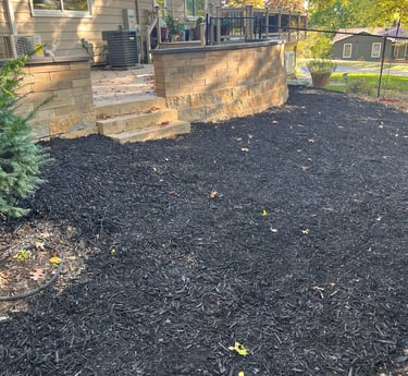 Residential backyard featuring a stone retaining wall and patio surrounded by fresh black mulch.