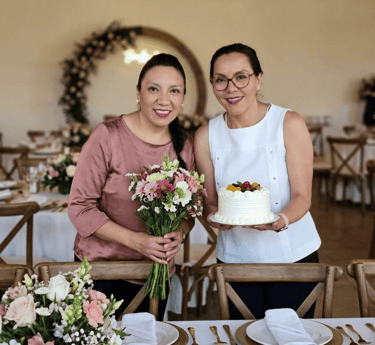 mujeres sosteniendo una flores y un pastel detrás de una mesa montada para un evento