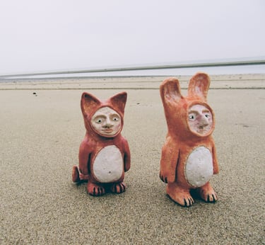 two small ceramic animals sitting on a sandy beach