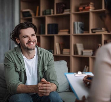 A smiling man sits on a sofa during a therapy session with a mental health professional.