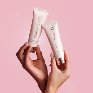 The photo shows two hands holding a HighOnLove product  - a hand cream, in front of a light pink background..
