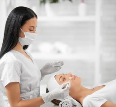 Professional esthetician in a mask applying a chemical peel facial treatment to a client in a spa.