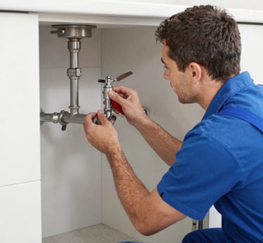 A cleaning cart is positioned in an outdoor area with a concrete building structure in the background. The cart holds a large bucket and cleaning supplies, including a striped cloth and a pink cloth hanging over the handle. A green scrubbing brush is placed on the bottom platform. Surrounding the area are vibrant flower beds with yellow and white flowers, as well as bushes and trees, contributing to a tidy and well-maintained environment.