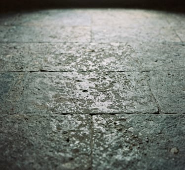 Moody heritage photography of a restored stone floor in an old stable, soft natural window light filtering through a small opening, desaturated stone grays, 35mm film grain, close-up texture.