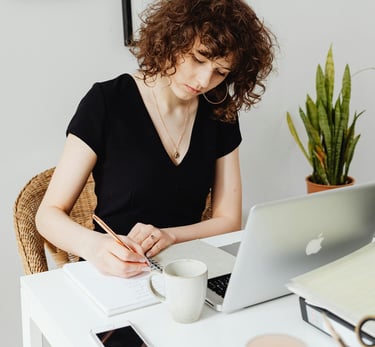 A woman in a black top sitting in front of an open laptop and wrting on a notepad