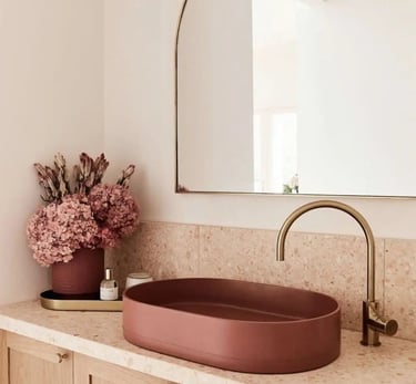 Banheiro elegante com cuba redonda rosé sobre bancada clara, torneira dourada, espelho orgânico e de
