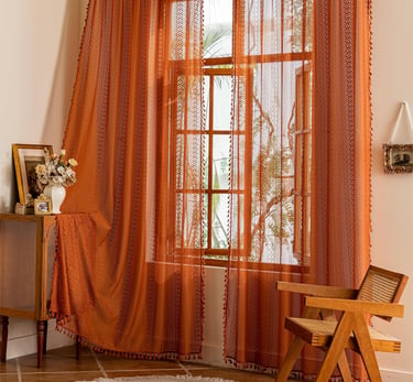 Burnt orange boho sheer curtains with tassels filter sunlight in a rustic living room window.
