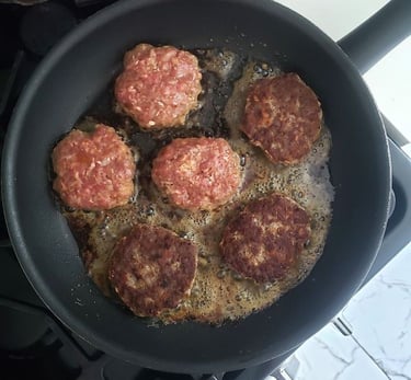 Sizzling ground beef burger patties frying in a non-stick skillet on a gas stovetop.